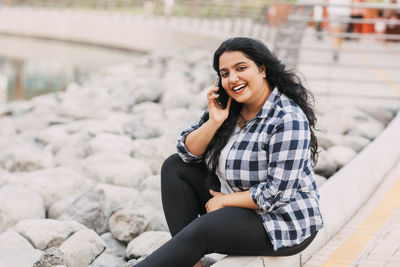 Portrait of a young indian woman talking on the phone, sitting on the embankment