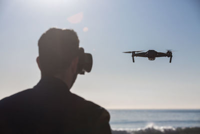 Back view of unrecognizable male wearing modern vr goggles operating drone with remote controller and experiencing virtual reality while standing against sea