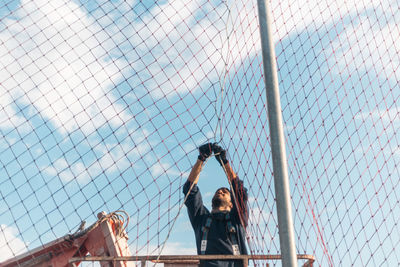 Low angle view of people on chainlink fence against sky