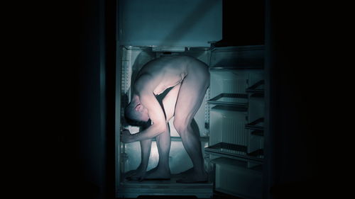 Full length of naked man standing in refrigerator in darkroom