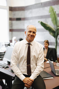 Portrait of smiling senior male lawyer sitting on desk in office