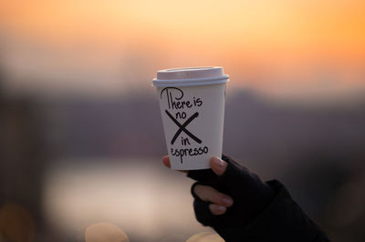 Cropped image of person holding disposable coffee cup with text during sunset