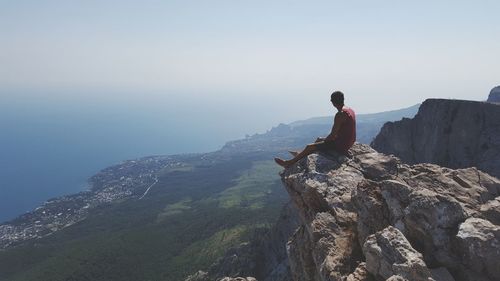 Rear view of man sitting on rock by sea against clear sky
