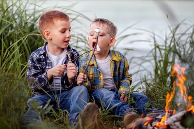 Siblings eating food while sitting by campfire