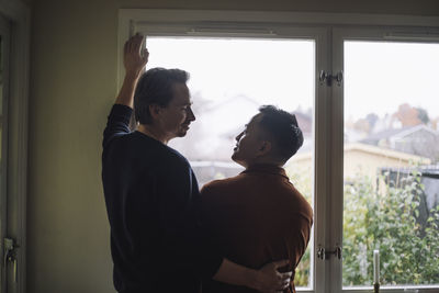 Rear view of multiracial gay couple talking while standing near window at home