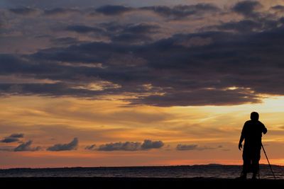 Rear view of silhouette man with tripod standing at beach during sunset