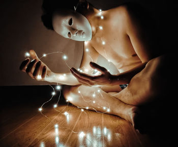 Shirtless young man with string lights wearing mask while sitting in darkroom