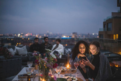 Smiling woman showing smart phone to female while friends sitting in background during social gathering on building terr