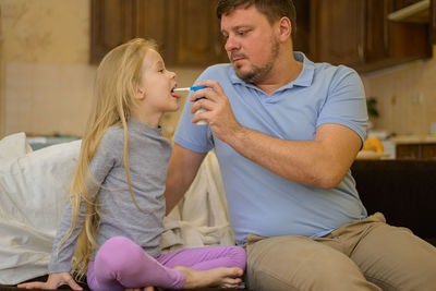 Dad squirts spray medicine down the throat of his sick child on the couch at home. the father