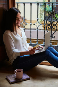 Young woman shopping and paying online on a laptop with credit card while relaxing at home next to a window.