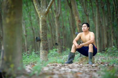 Full length of thoughtful shirtless man sitting in forest