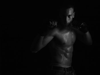 Shirtless young man boxing while standing against black background