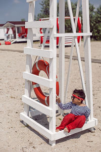 Child boy in striped clothes and red pants  standing on beach. white lifeguard tower, with a circle