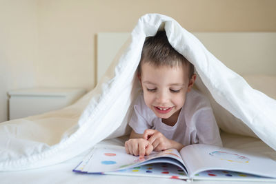 Adorable smiling kid lying down on bed under blanket, reading a book