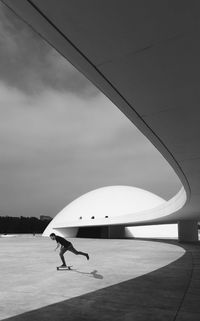 Black and white of unrecognizable male skater performing trick on skateboard on pavement near contemporary building with futuristic architecture