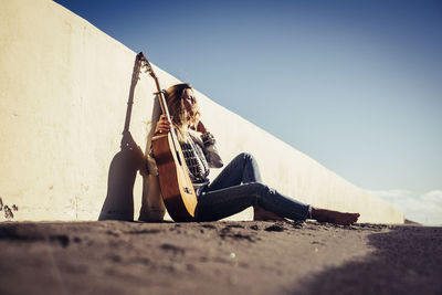Full length of young woman with guitar leaning on retaining wall against clear sky