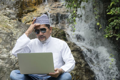 Mature man wearing sunglasses while using laptop against waterfall