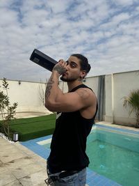 Side view of young man drinking water while standing against swimming pool