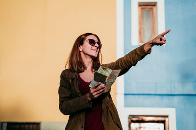 Smiling woman with map standing against wall during sunny day