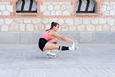 Full body side view of active hispanic female in roller skates doing stunt while riding on one leg on paved sidewalk near building