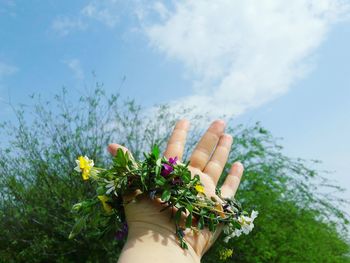 Cropped hand of person holding floral wreath against sky