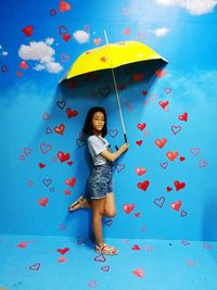 Portrait of young girl holding umbrella while standing against blue wall with heart shape