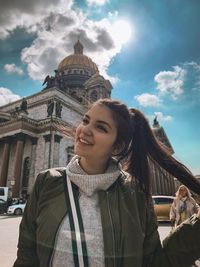 Portrait of smiling young woman with building against cloudy sky