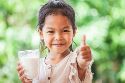 Portrait of girl gesturing thumbs up sign while holding milk glass outdoors