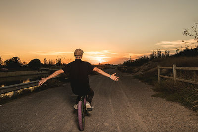 Man with arms outstretched riding bicycle against sky during sunset