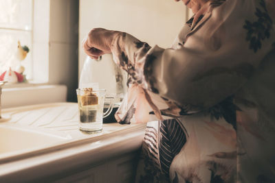 Cropped unrecognizable adult female wearing silk blouse and trousers brewing tea bag in glass mug kitchen