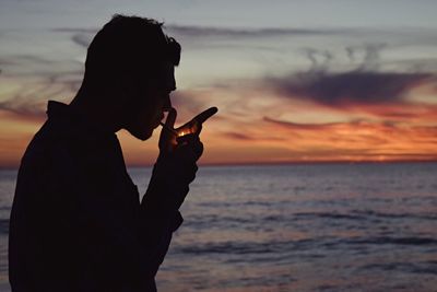 Silhouette man lighting cigarette by sea during sunset