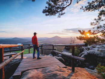 Rear view of man standing on railing against sky during sunset