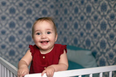 Cheerful smiling baby girl in red overall standing at the railing of her bed.