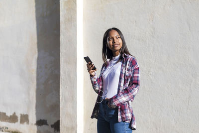 Young woman using phone while standing against wall