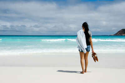 Rear view of mid adult woman standing at beach against cloudy sky
