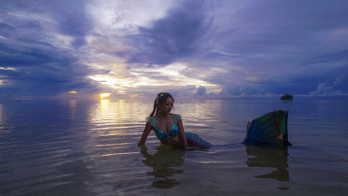 Female model in mermaid costume posing on shore against cloudy sky
