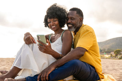 Side view of man using mobile phone while sitting on sand at beach