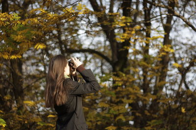 Side view of young woman photographing while standing against trees in forest