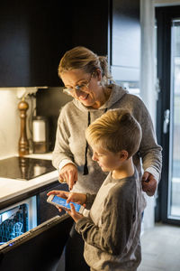Mother guiding son while using mobile app over dishwasher in kitchen at home