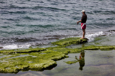Mature man fishing while standing on moss covered rock by sea