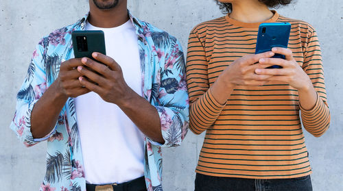 Cropped unrecognizable multiracial couple of partners in trendy clothes with afro hairstyle standing near wall with cellphones