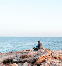 Man sitting on rock looking at sea against clear sky