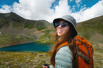 Beautiful young woman with a tourist backpack in a sports jacket and hat. summer landscape of lake