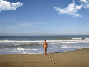 Rear view of naked man standing on shore at beach against sky