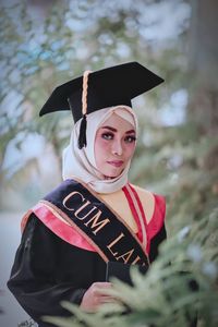 Portrait of woman wearing graduation gown while standing against tree