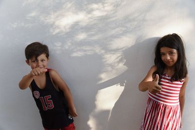 Portrait of siblings showing thumbs up while standing against white wall
