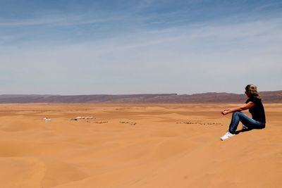 Side view of woman sitting on sand dune at sahara desert against sky