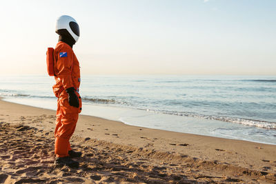 Side view of astronaut in futuristic spacesuit and helmet standing on sand and admiring waving sea while spending time on beach in evening