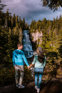 Rear view of people standing against waterfall and trees in forest