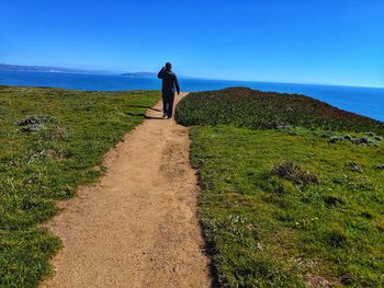 Rear view of man walking on dirt road against sea and sky